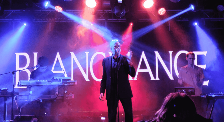 A band performs on a stage with dramatic blue and purple lighting. The lead singer stands at the center with a microphone, flanked by two musicians on keyboards. “BLANCMANGE” is displayed in large letters, reminiscent of a hot 1027 breakfast show backdrop.