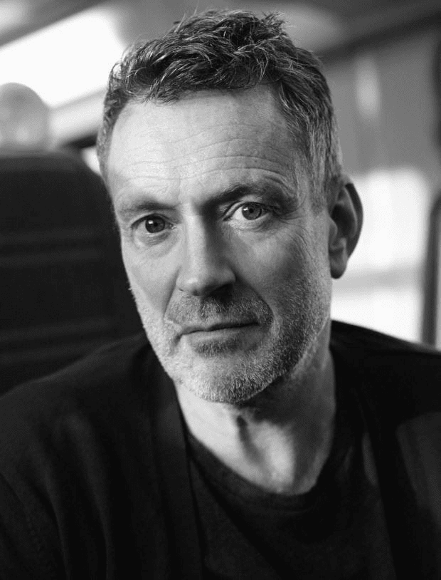 Black and white portrait of a middle-aged man with short, slightly curly hair, a trimmed beard, and a serious expression, sitting indoors—perhaps awaiting an interview on Hot 1027 Breakfast—with blurred seats in the background.