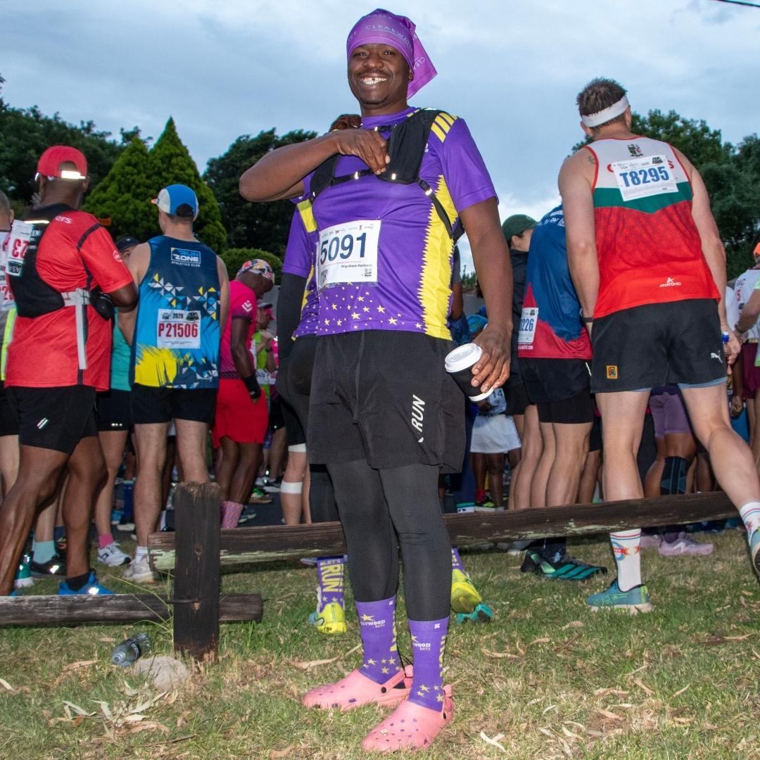 A smiling runner in a purple outfit and pink Crocs, perhaps remembering Last Christmas, stands in front of a crowd at a race event, holding a small cup and wearing bib number 5091. Grass and trees can be seen in the background.