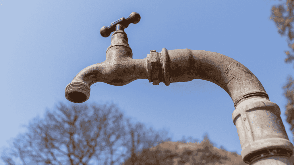 A close-up of an old, weathered water tap with no handle, set against a clear blue sky and blurred trees in the background, evokes the rustic aesthetic often featured in Die Kantoor.