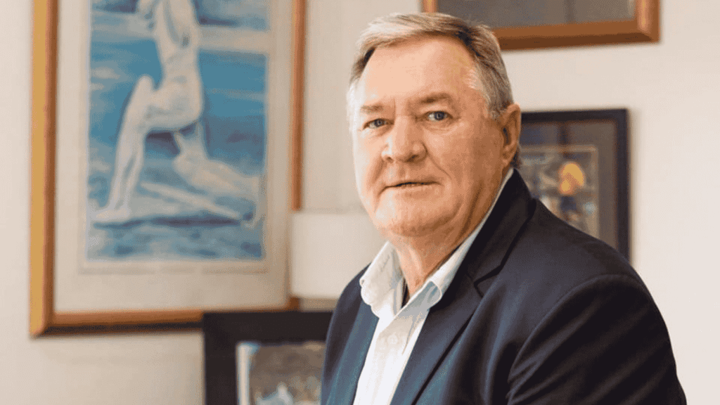 An older man with short gray hair and a light blue shirt under a dark blazer sits indoors, looking at the camera. Behind him, framed artwork and paintings give the space a warm, inviting vibe—perfect for tuning in to Hot 1027 Breakfast.