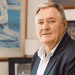 An older man with short gray hair and a light blue shirt under a dark blazer sits indoors, looking at the camera. Behind him, framed artwork and paintings give the space a warm, inviting vibe—perfect for tuning in to Hot 1027 Breakfast.