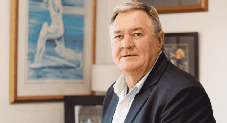 An older man with short gray hair and a light blue shirt under a dark blazer sits indoors, looking at the camera. Behind him, framed artwork and paintings give the space a warm, inviting vibe—perfect for tuning in to Hot 1027 Breakfast.