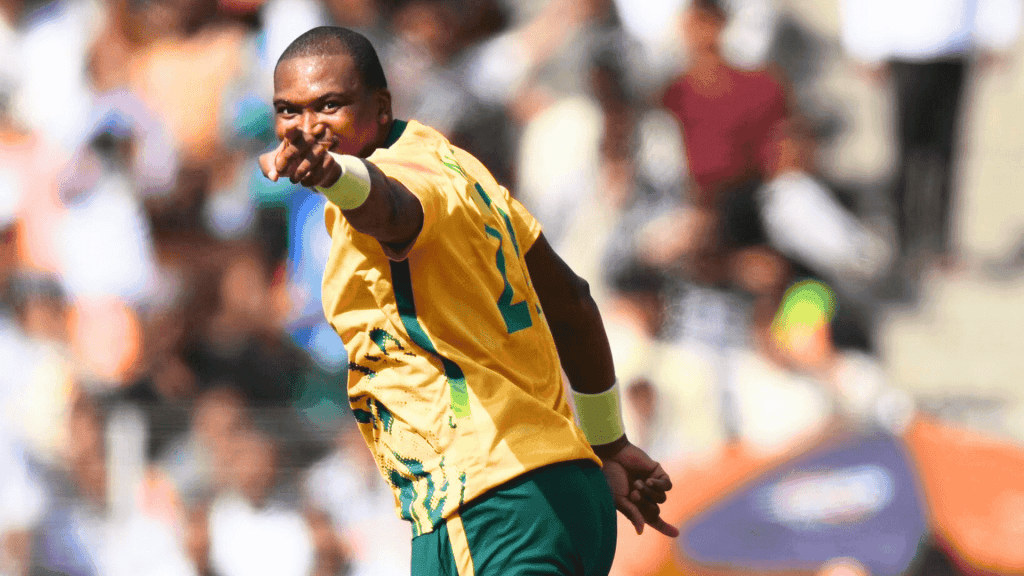 A cricket player in a yellow and green jersey points forward, celebrating on the field with a blurred crowd in the background, as vibrant as the energy of Hot 1027 Breakfast.