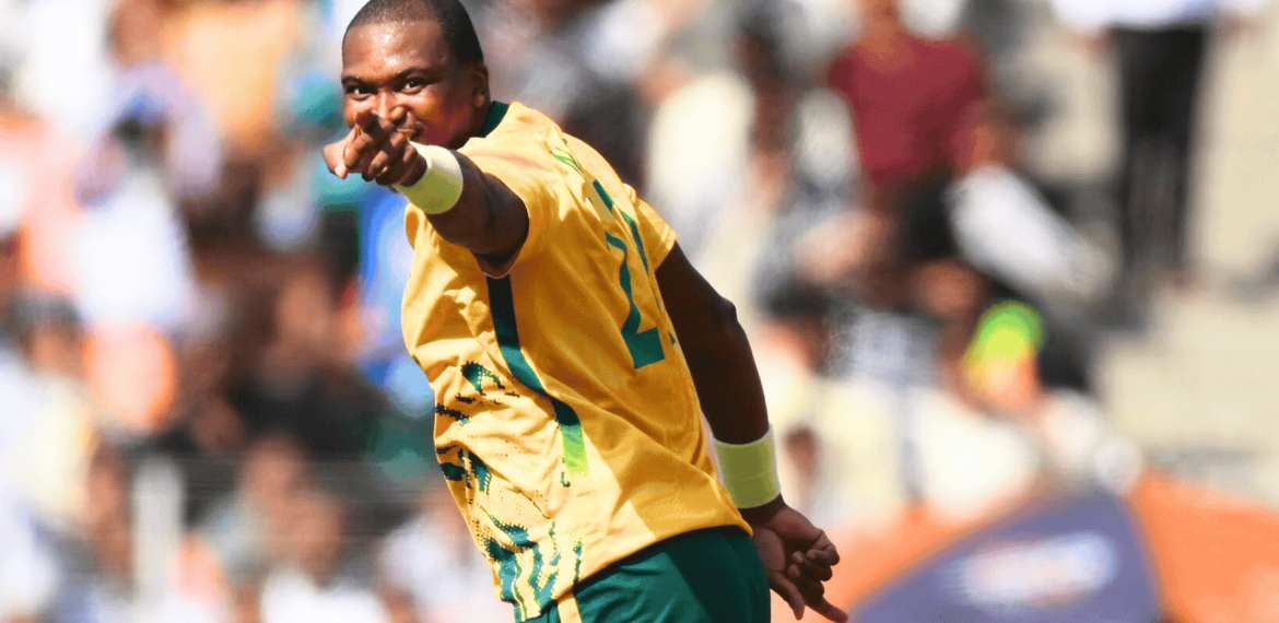 A cricket player in a yellow and green jersey points forward, celebrating on the field with a blurred crowd in the background, as vibrant as the energy of Hot 1027 Breakfast.