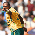 A cricket player in a yellow and green jersey points forward, celebrating on the field with a blurred crowd in the background, as vibrant as the energy of Hot 1027 Breakfast.