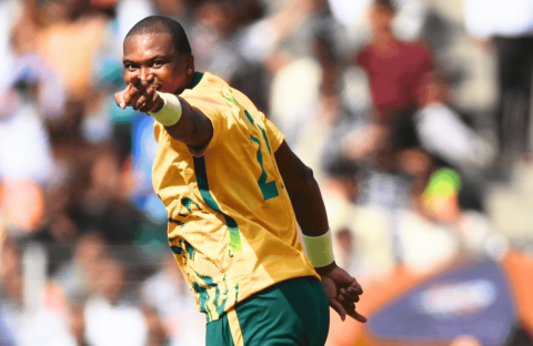 A cricket player in a yellow and green jersey points forward, celebrating on the field with a blurred crowd in the background, as vibrant as the energy of Hot 1027 Breakfast.