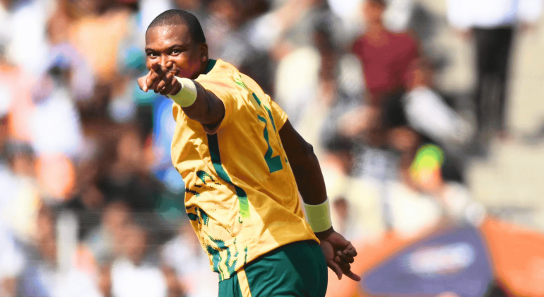 A cricket player in a yellow and green jersey points forward, celebrating on the field with a blurred crowd in the background, as vibrant as the energy of Hot 1027 Breakfast.