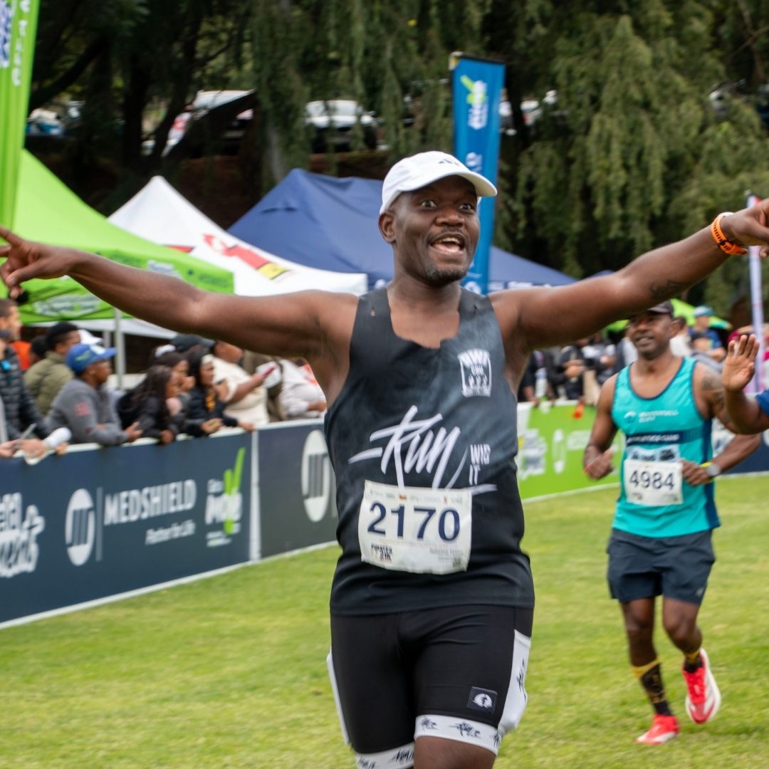 A runner wearing a black tank top, white cap, and race bib 2170 smiles with arms outstretched as he nears the finish line—just like he did last Christmas. Spectators and another runner cheer in the background.