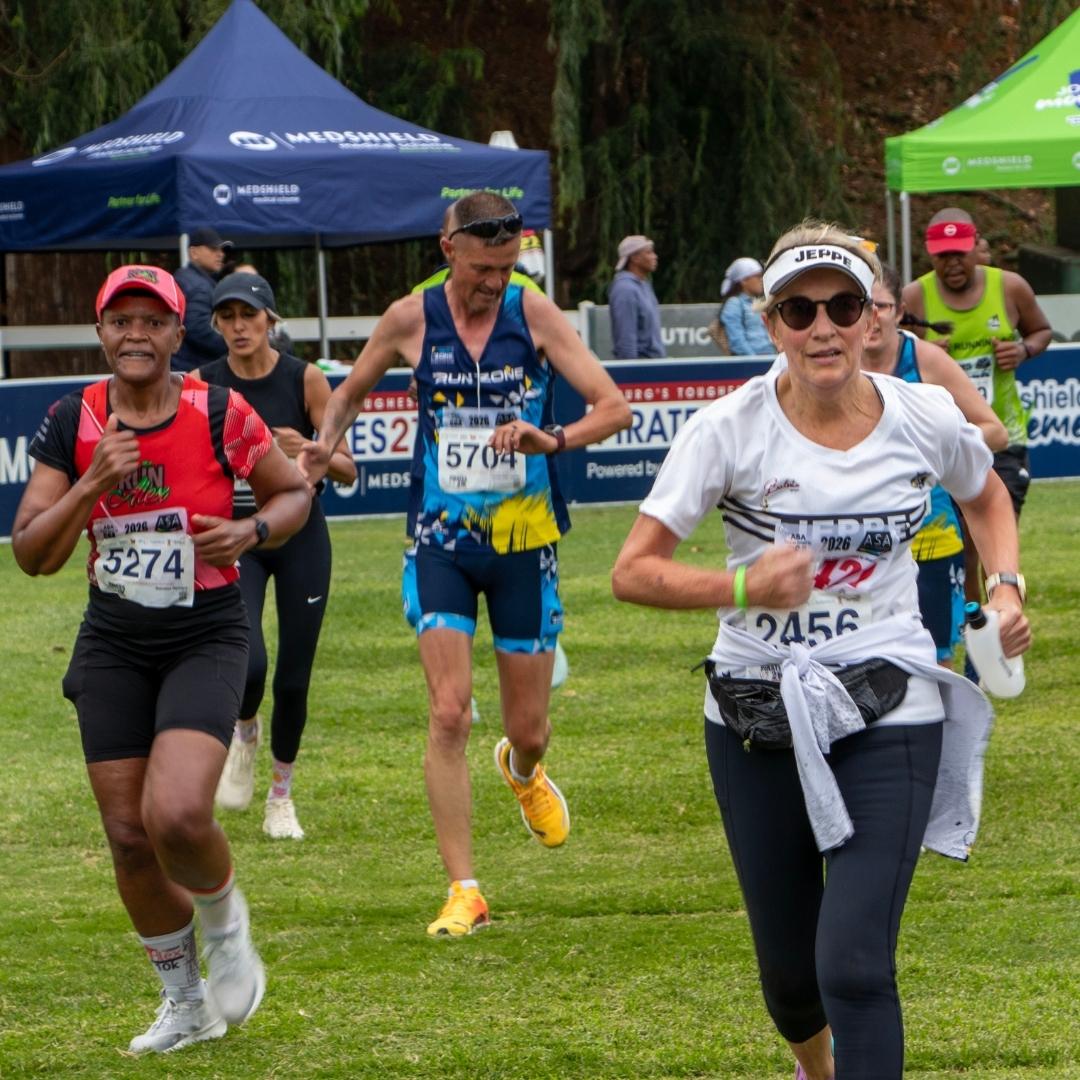 A group of marathon runners, wearing numbered bibs and athletic gear, dash on grass past tents and banners. The front runner in sunglasses and a white shirt leads the pack—perhaps chasing the spirit of Last Christmas with every stride.