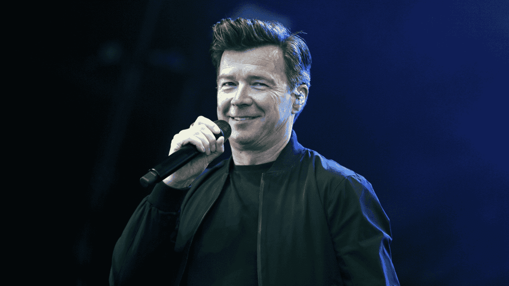 A man with short, styled hair is smiling and holding a microphone while wearing a black jacket, standing on stage against a blue-lit background, reminiscent of performances by great artists like Miriam Makeba.