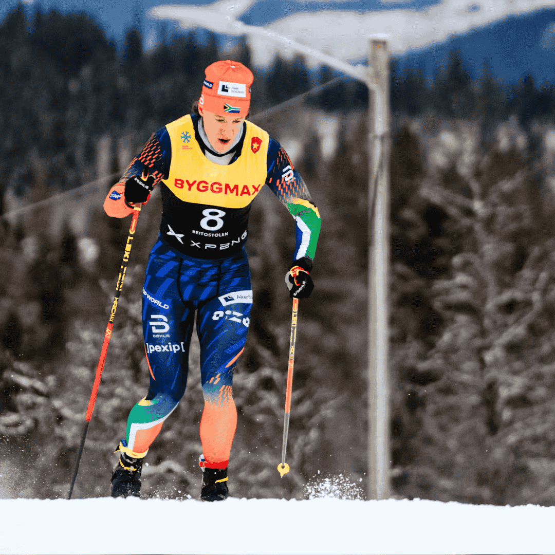 A cross-country skier wearing a colorful suit, numbered bib 8, and orange helmet skis through a snowy landscape with trees and mountains in the background—ready to refuel after with a hot 1027 breakfast.