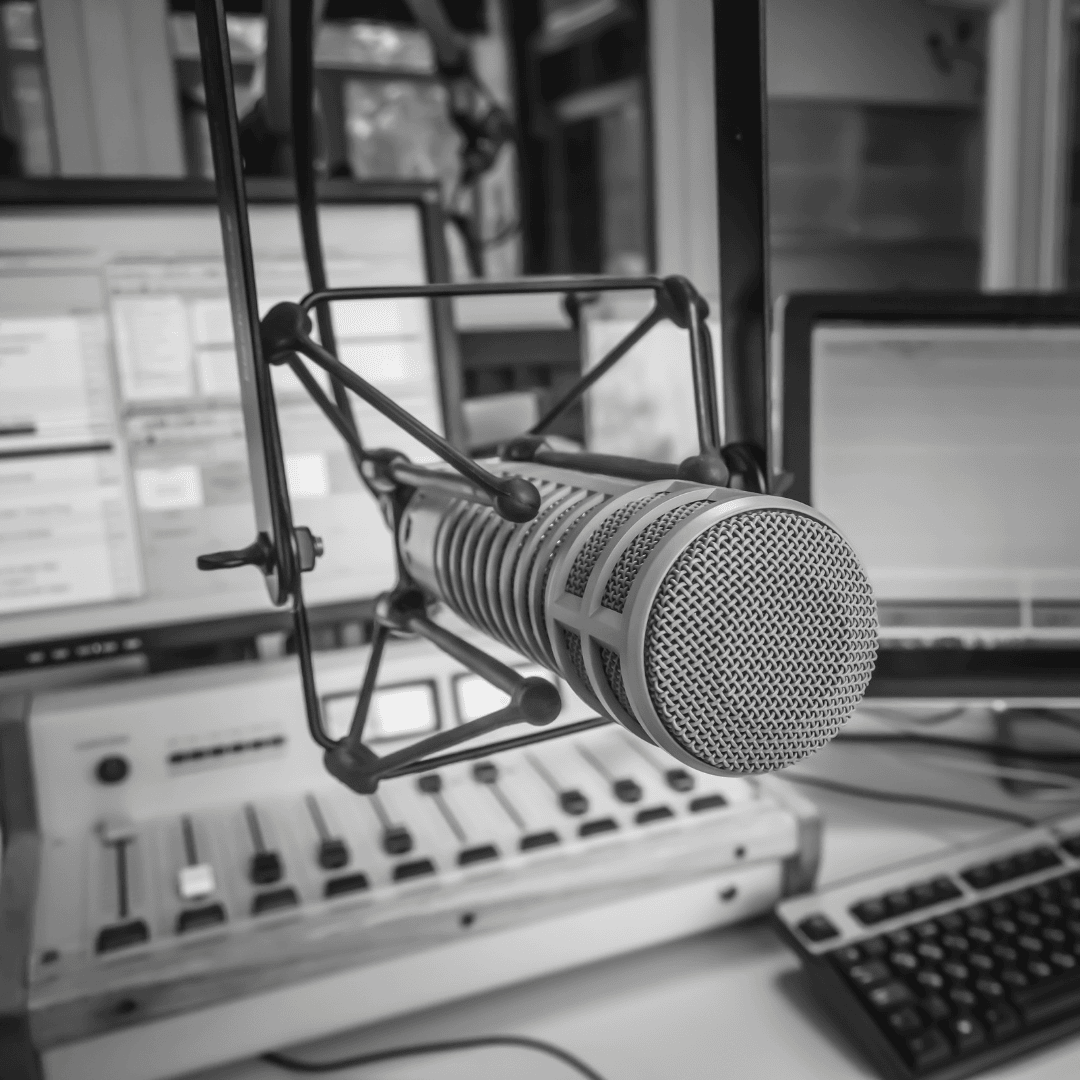 A large microphone on a suspension mount is positioned in front of a mixing console, computer monitors, and a keyboard in a radio broadcasting studio—evoking the spirit of legends like Miriam Makeba. The image is in black and white.