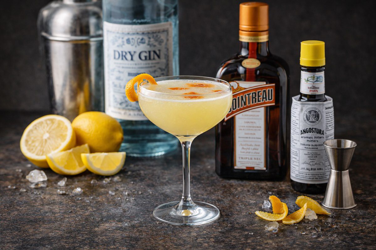 A yellow cocktail in a coupe glass garnished with orange peel sits on a counter surrounded by lemons, a gin bottle, Cointreau, Angostura bitters, a jigger, shaker, and ice cubes at this dog friendly bar.