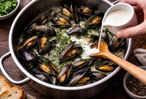 A pot of cooked mussels garnished with chopped herbs, with a hand pouring cream and a wooden spoon stirring. Slices of bread and bowls of herbs and pepper are nearby in this cozy, dog friendly setting.