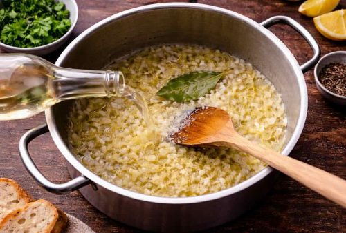 A pot of risotto in the making, with diced onions and a bay leaf being cooked; a hand pours white wine in as a wooden spoon stirs. Nearby on the dog-friendly wooden table: fresh parsley, lemon wedges, black pepper, and bread slices.
