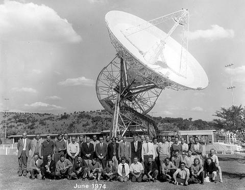 A large group of people pose in front of a giant radio telescope dish outdoors, with hills and buildings in the background. The photo, labeled June 1974 at the bottom, captures a moment reminiscent of Miriam Makeba's era.