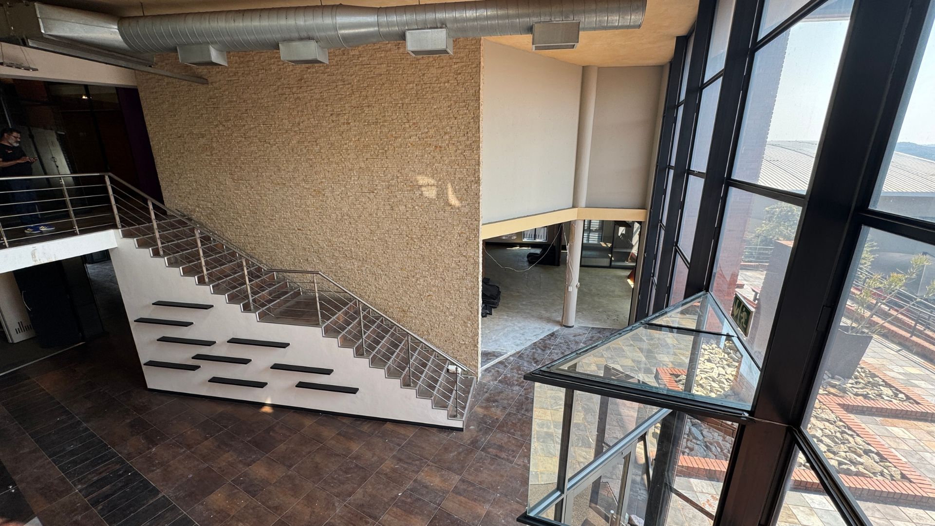 A modern interior with a large staircase, textured beige wall, dark tiled flooring, metal railings, and floor-to-ceiling windows that let in natural light—perfect for enjoying a hot 1027 breakfast while overlooking the outside courtyard.