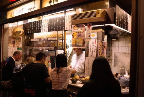 A small, busy Japanese ramen shop offers the perfect travel getaway, with diners seated at the counter enjoying food. Steam rises from the kitchen where a chef cooks, surrounded by menus written in Japanese on the walls.