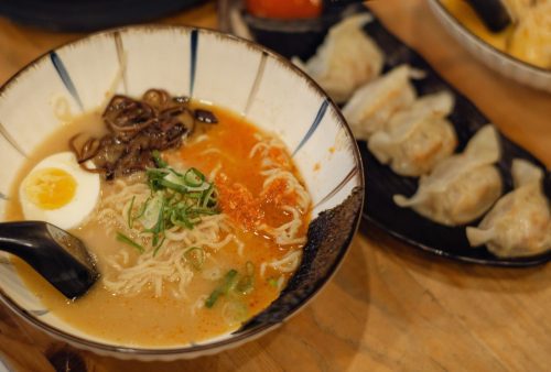 A bowl of ramen with sliced green onions, half a boiled egg, mushrooms, and red seasoning sits next to gyoza dumplings on a wooden table—a perfect travel-inspired getaway meal for adventurous food lovers.