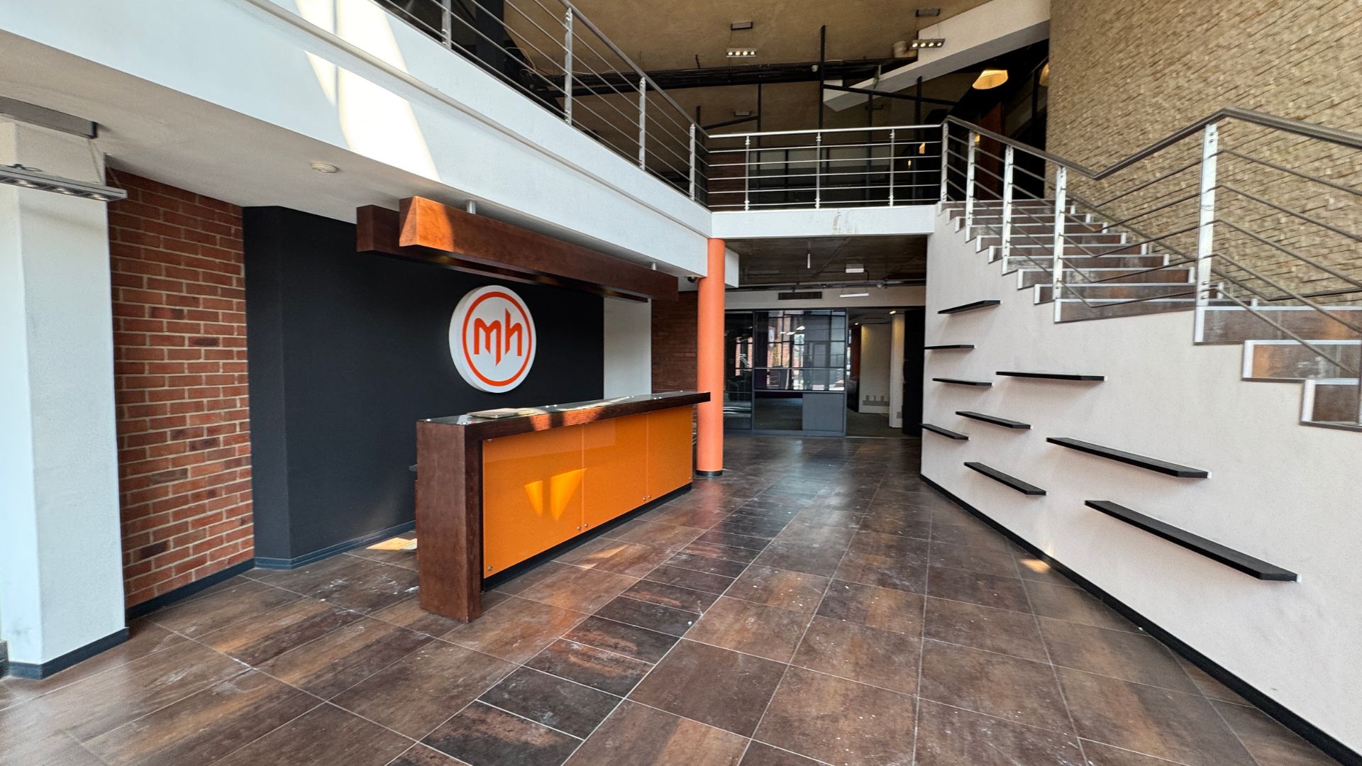 Modern office lobby with a brown tile floor, orange reception desk, black accent wall featuring a circular mh logo, and an open staircase. Exposed brick and white walls create a vibrant space, perfect for hosting a hot 1027 breakfast as natural light streams in.