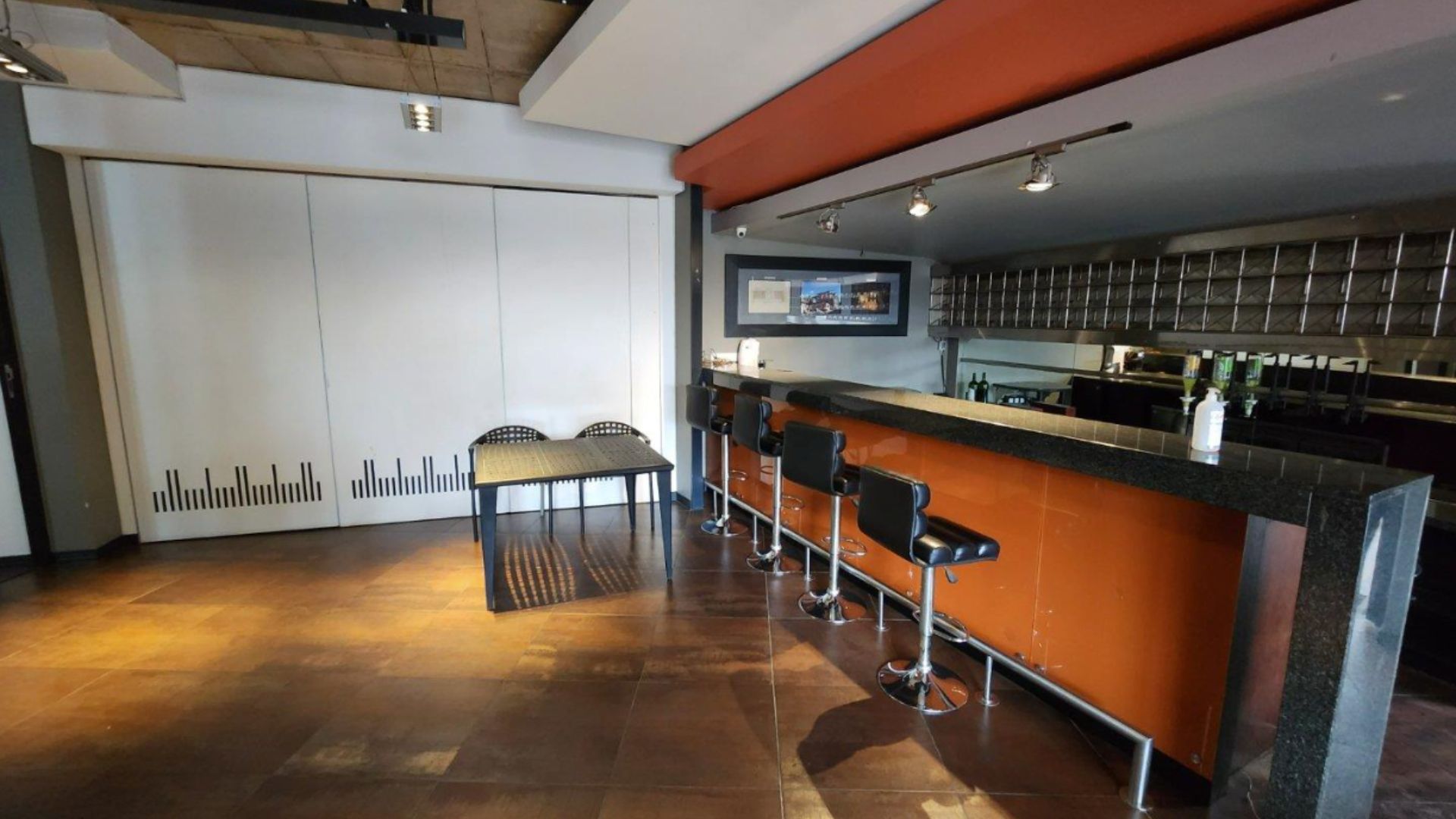 Modern kitchen with an orange and black bar counter, four black bar stools perfect for a hot 1027 breakfast, a black table with two checkered chairs, and a large white sliding door on the left. Ceiling lights and framed art above the bar.