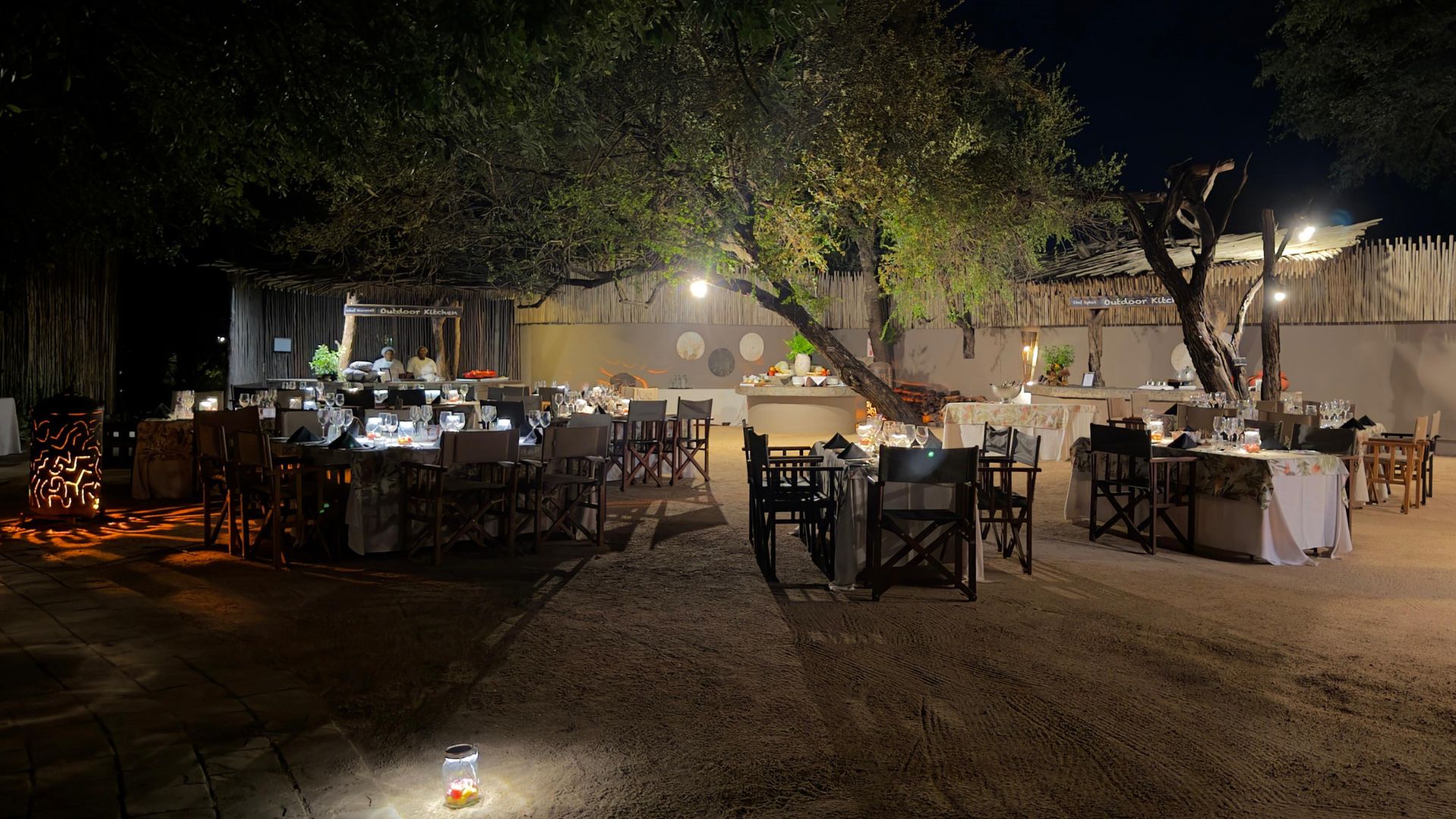 Outdoor evening dining setup with several tables and chairs arranged under large trees, illuminated by soft lights—perfect for a relaxing matric celebration. Food and decorations are displayed on buffet tables in the background, set on sandy ground.