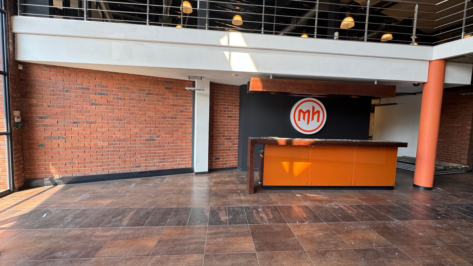 A modern lobby with a brick wall, a black and orange reception desk, and a circular mh logo on the wall. The space features large windows, tiled flooring, an upper balcony with railing, hanging lights, and a cozy hot 1027 breakfast corner.