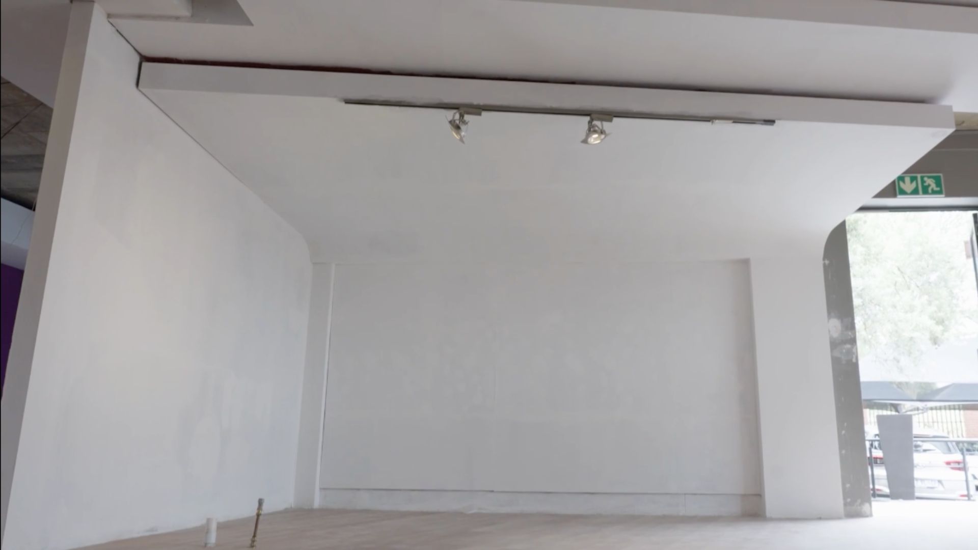 A mostly empty white room with two ceiling spotlights, white walls, and a light-colored floor. An emergency exit sign and some daylight are visible on the right side near a doorway—perfect for a hot 1027 breakfast event.