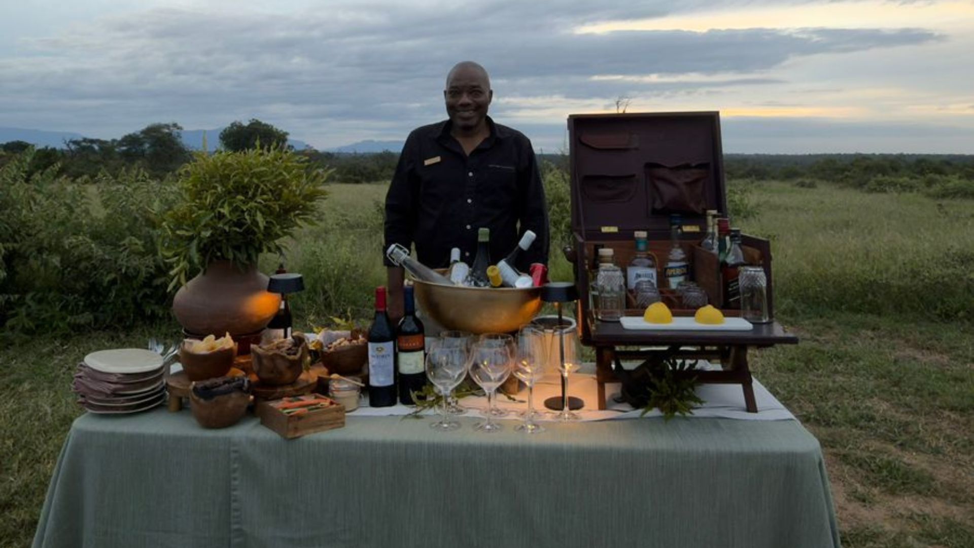 A smiling person stands behind an outdoor bar setup on a table, displaying wine, liquor bottles, glasses, snacks, and decorations, celebrating their Matric with grassy fields and a cloudy sky in the background.
