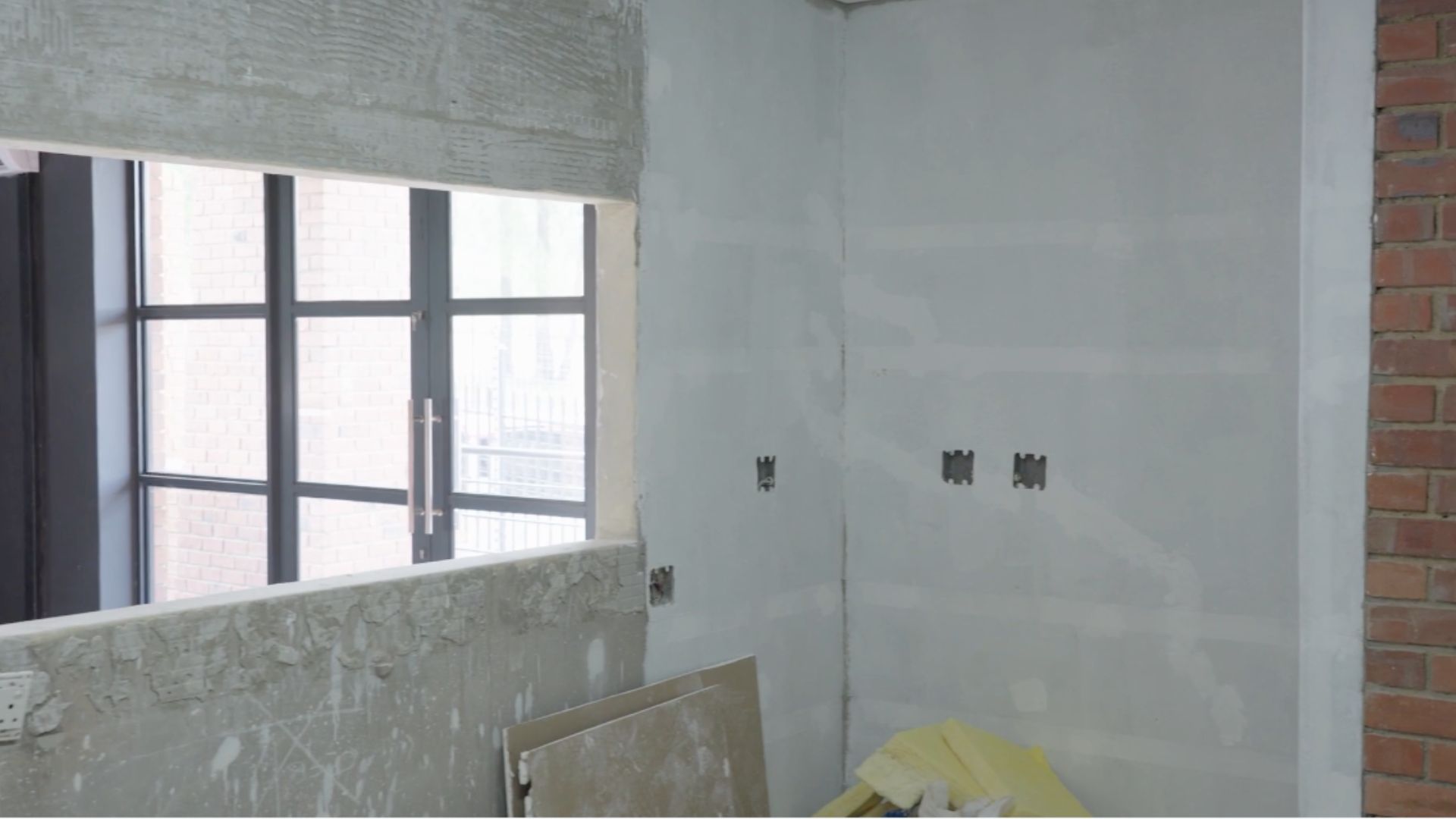 A room under construction with unfinished walls, exposed electrical outlets, a large window with black frames, and construction materials leaning against the wall—looks like it’s not ready for a hot 1027 breakfast just yet.