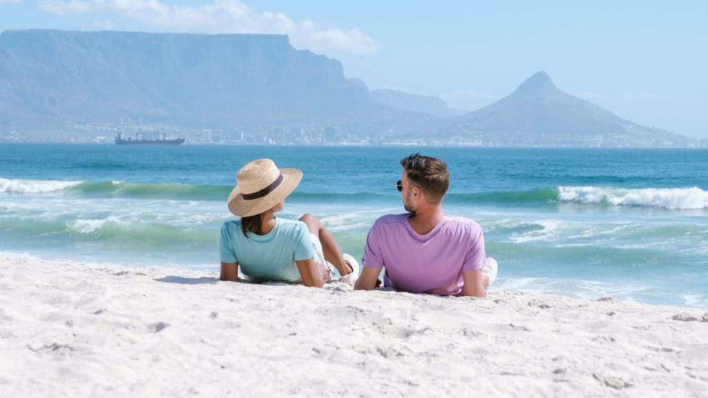 A couple relaxes on a sandy beach, enjoying their matric holiday as they face the ocean with mountains and a city skyline in the background. The woman wears a straw hat and light blue shirt, while the man wears sunglasses and a lavender shirt.