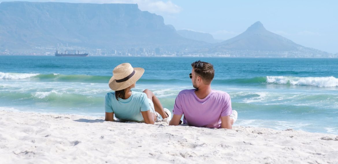 A couple relaxes on a sandy beach, enjoying their matric holiday as they face the ocean with mountains and a city skyline in the background. The woman wears a straw hat and light blue shirt, while the man wears sunglasses and a lavender shirt.