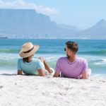 A couple relaxes on a sandy beach, enjoying their matric holiday as they face the ocean with mountains and a city skyline in the background. The woman wears a straw hat and light blue shirt, while the man wears sunglasses and a lavender shirt.