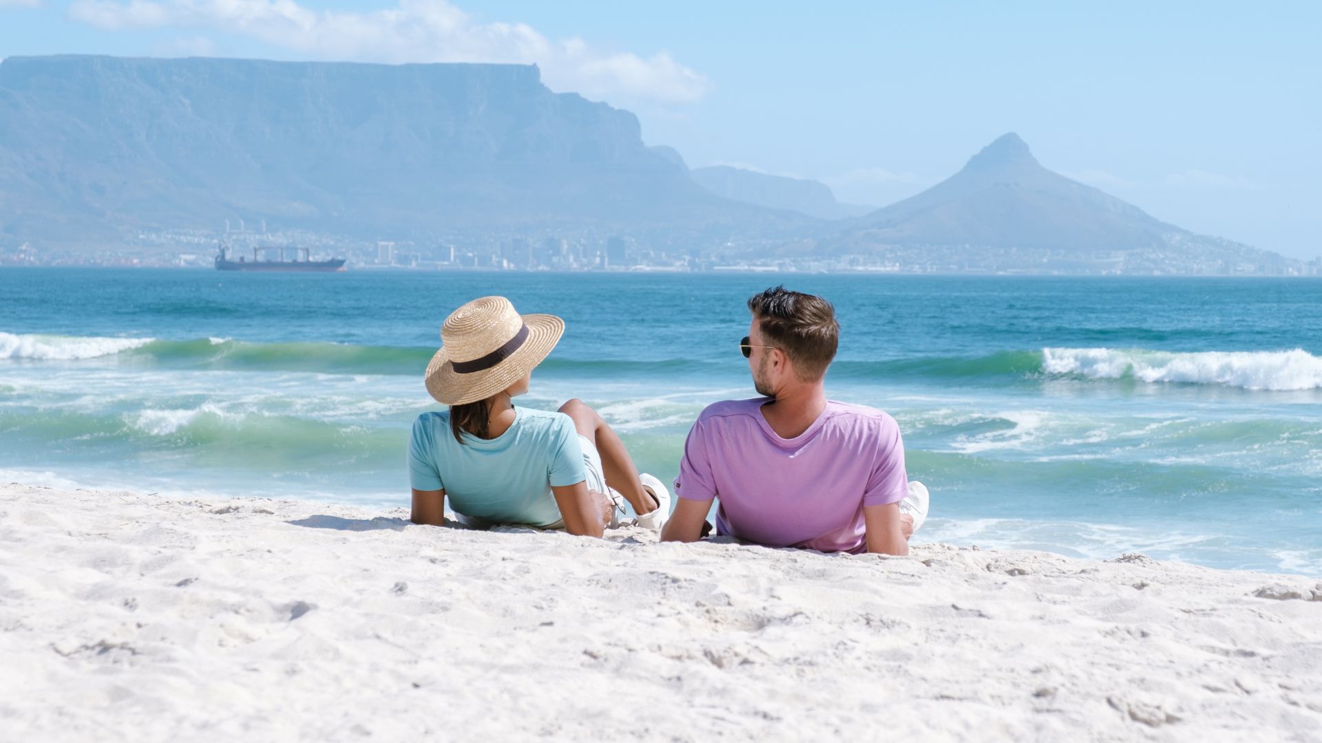 A couple relaxes on a sandy beach, enjoying their matric holiday as they face the ocean with mountains and a city skyline in the background. The woman wears a straw hat and light blue shirt, while the man wears sunglasses and a lavender shirt.