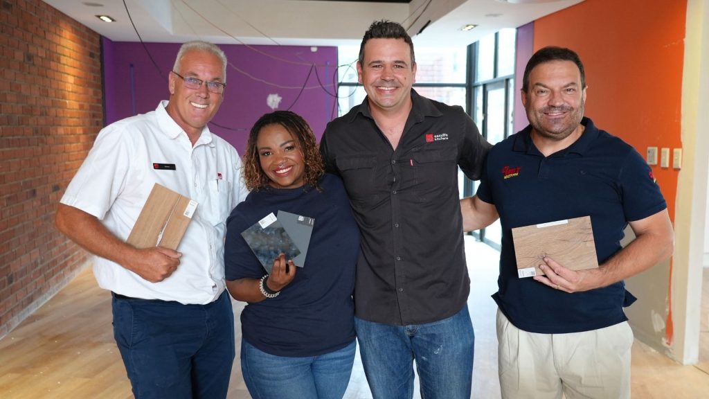 Four people stand indoors, smiling at the camera. Three of them hold different flooring samples. The room has red brick and orange painted walls, with some construction wires and unfinished surfaces—a real hot 1027 breakfast vibe.