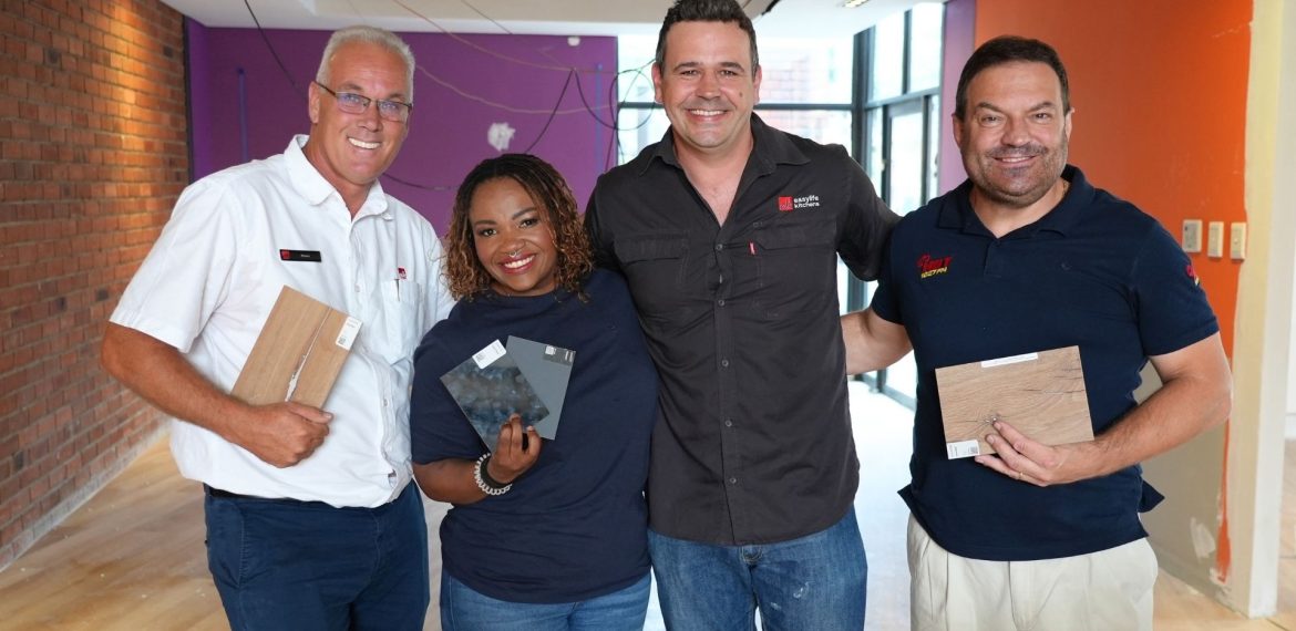 Four people stand indoors, smiling at the camera. Three of them hold different flooring samples. The room has red brick and orange painted walls, with some construction wires and unfinished surfaces—a real hot 1027 breakfast vibe.
