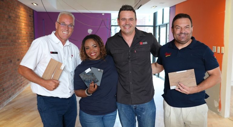 Four people stand indoors, smiling at the camera. Three of them hold different flooring samples. The room has red brick and orange painted walls, with some construction wires and unfinished surfaces—a real hot 1027 breakfast vibe.