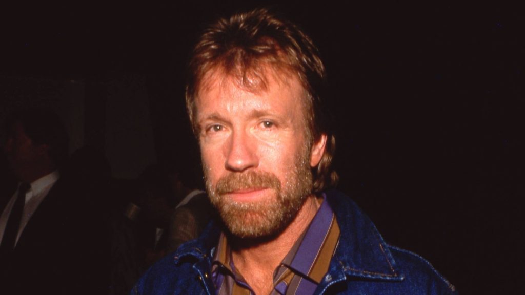 A man with light brown hair and a beard, wearing a blue jacket over a striped collared shirt, stands against a dark background, exuding the poised confidence of a recent Matric graduate.