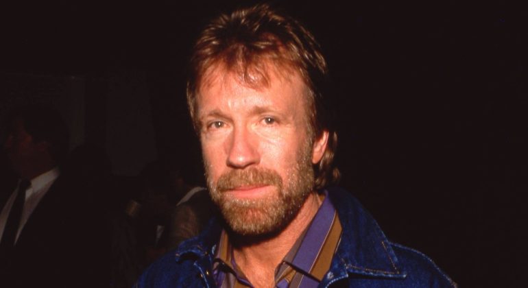 A man with light brown hair and a beard, wearing a blue jacket over a striped collared shirt, stands against a dark background, exuding the poised confidence of a recent Matric graduate.