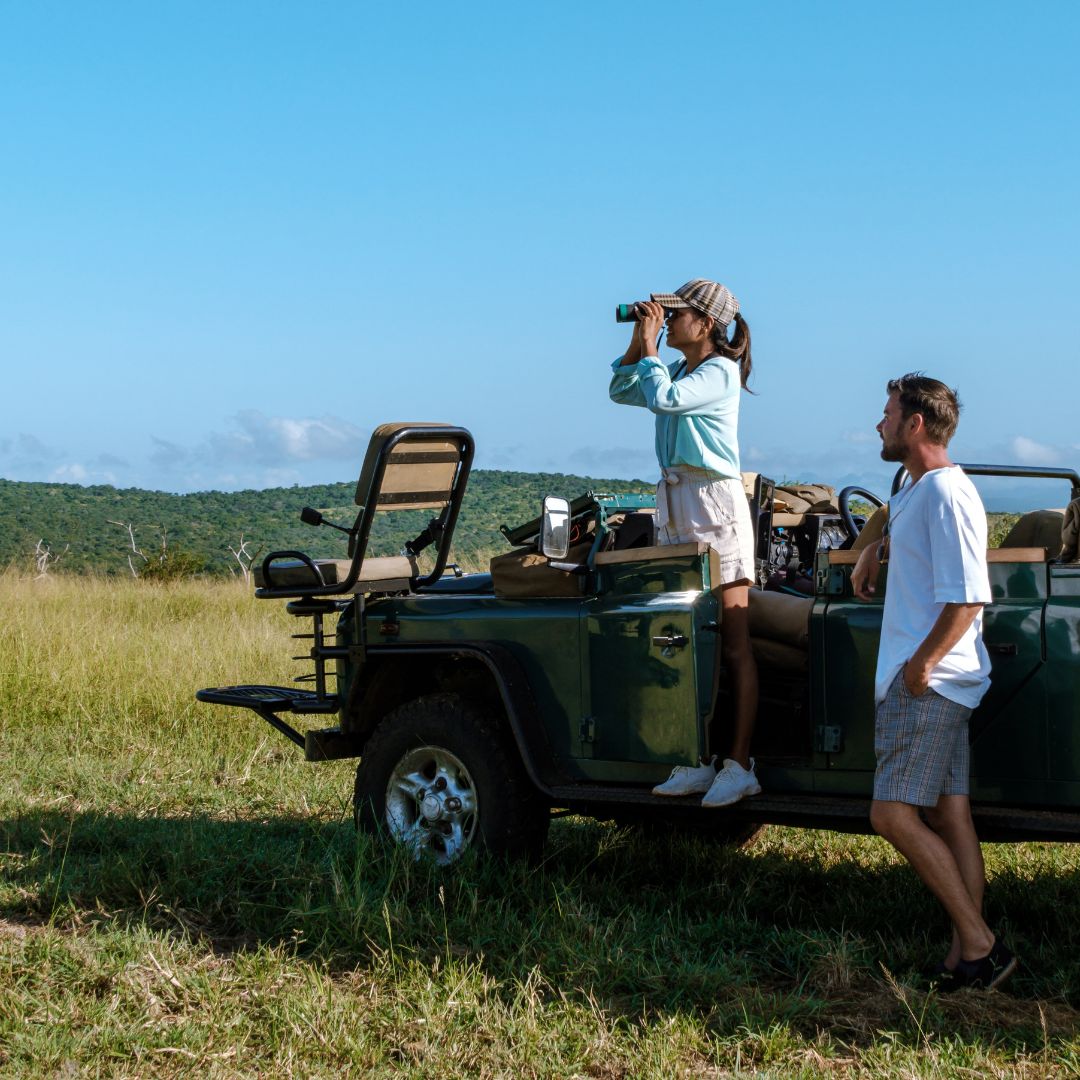 A woman, dressed in her Matric adventure gear, stands on the side step of a green safari jeep using binoculars, while a man stands nearby in a grassy savanna landscape under a clear blue sky.