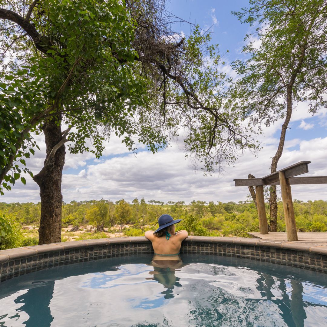 A person wearing a blue hat relaxes in a round outdoor pool after finishing Matric, surrounded by trees and lush greenery under a partly cloudy sky. Their back is to the camera, facing the scenic landscape.
