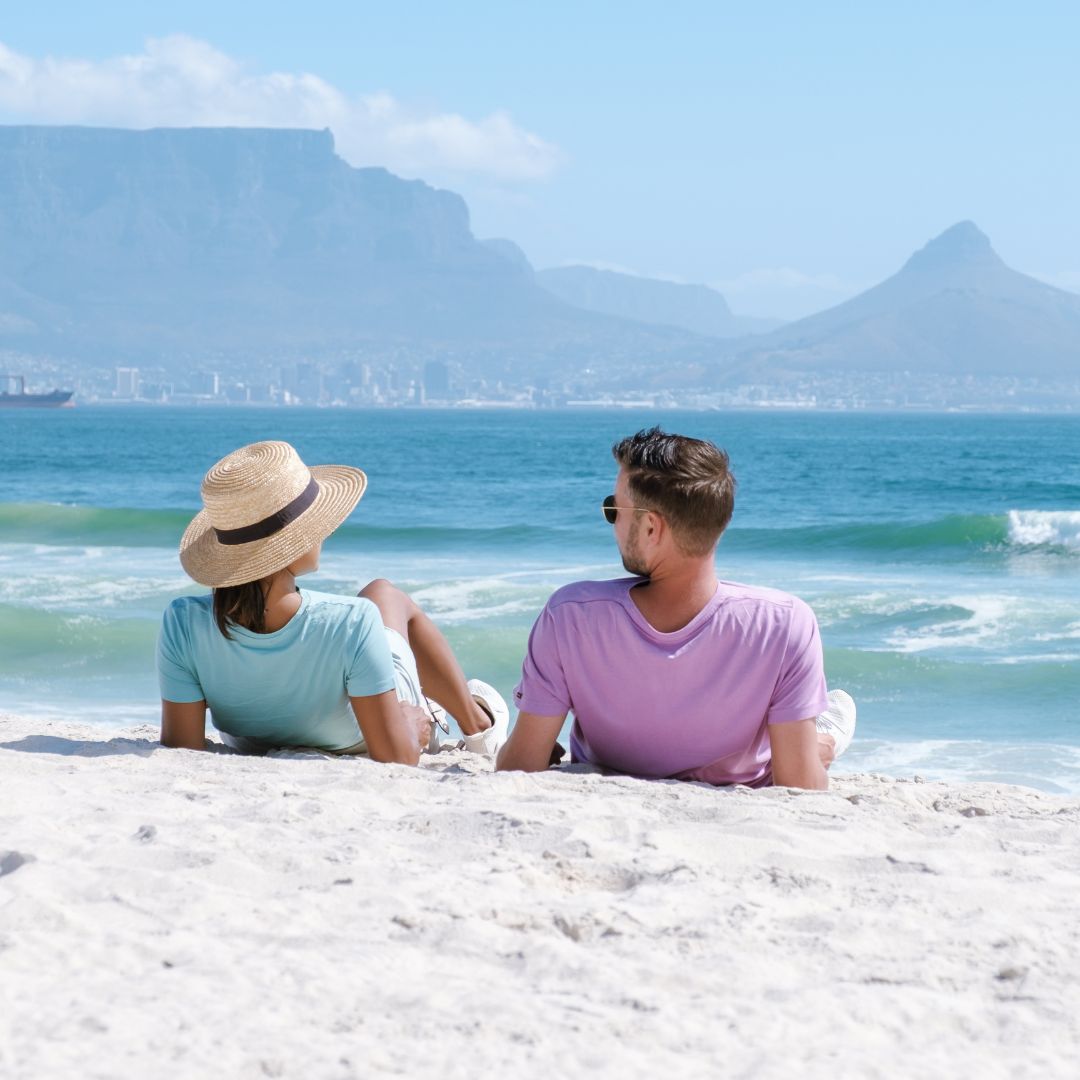 A couple relaxes on a sandy beach facing the ocean, with mountains and a city in the background, celebrating after Matric. The woman wears a sunhat and light blue shirt; the man wears sunglasses and a light purple shirt.