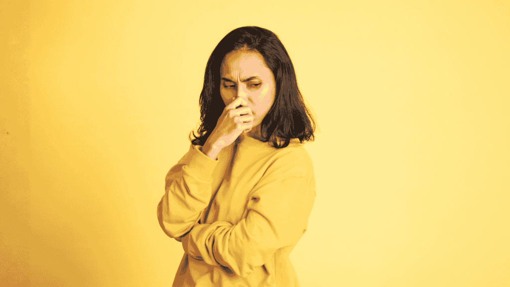 A woman in a yellow sweater stands against a yellow background, looking down with a worried or thoughtful expression—just like you might feel before coffee on the Hot 1027 Breakfast show—with her hand covering her mouth and her other arm crossed.