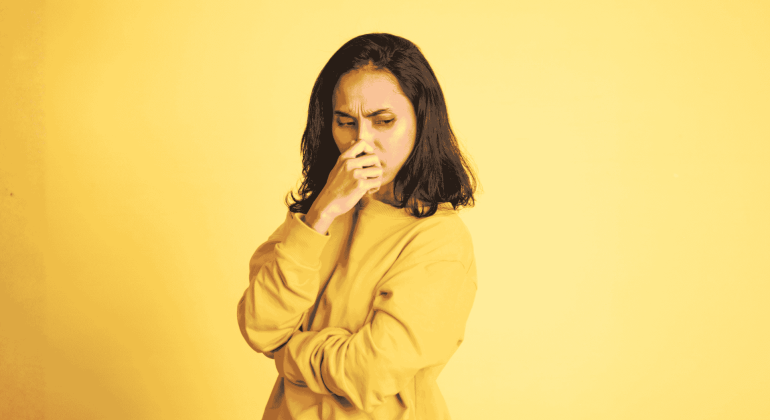 A woman in a yellow sweater stands against a yellow background, looking down with a worried or thoughtful expression—just like you might feel before coffee on the Hot 1027 Breakfast show—with her hand covering her mouth and her other arm crossed.