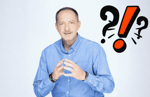 A man in a blue shirt gestures with his hands and smiles at the camera. Behind him are bold question marks and an exclamation mark, suggesting travel confusion or surprise. The background is light and plain.