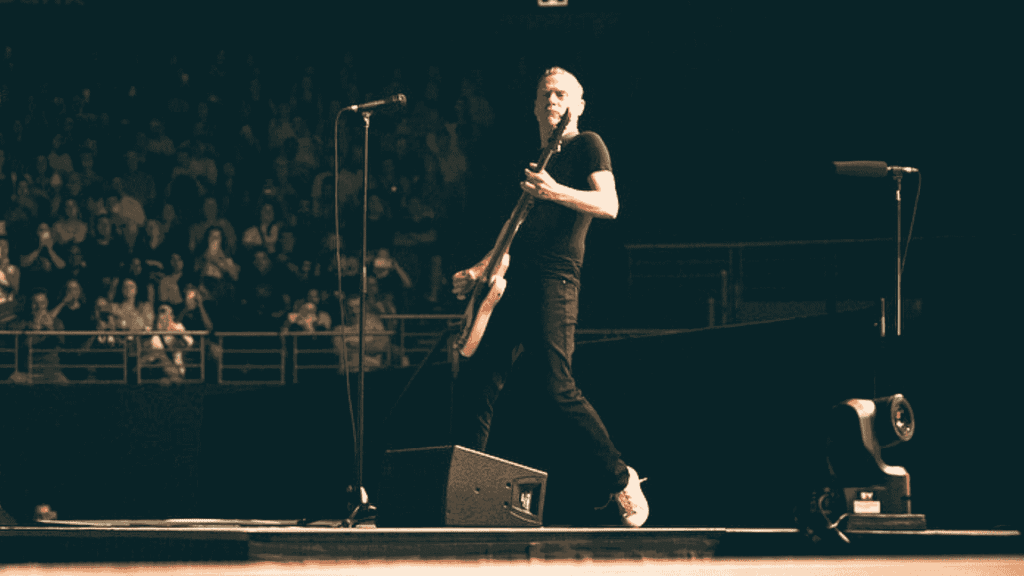 A musician in a black shirt and jeans plays an electric guitar on stage during a live Hot 1027 Breakfast performance, with a standing microphone nearby and a crowd of people watching in the background.