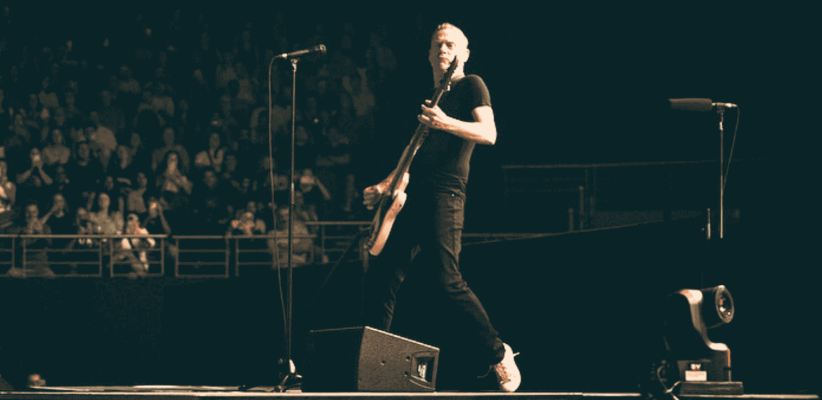 A musician in a black shirt and jeans plays an electric guitar on stage during a live Hot 1027 Breakfast performance, with a standing microphone nearby and a crowd of people watching in the background.