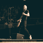 A musician in a black shirt and jeans plays an electric guitar on stage during a live Hot 1027 Breakfast performance, with a standing microphone nearby and a crowd of people watching in the background.
