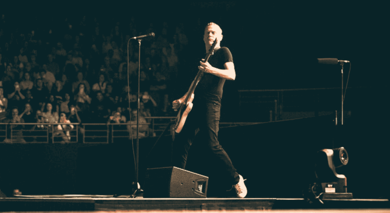 A musician in a black shirt and jeans plays an electric guitar on stage during a live Hot 1027 Breakfast performance, with a standing microphone nearby and a crowd of people watching in the background.